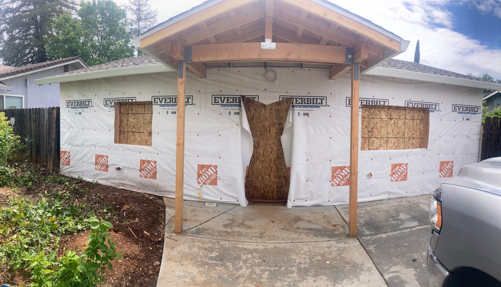 New gabled front porch cover with exposed wood beams, posts and metal connectors, beside house-wrapped walls and boarded openings during exterior renovation.