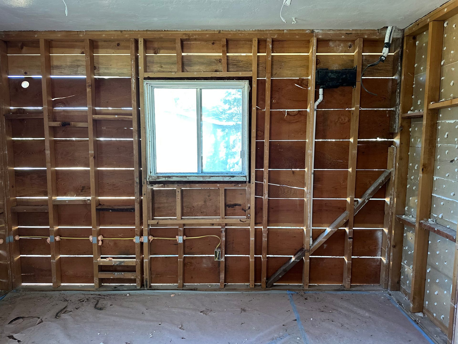 Stripped residential wall with original wood studs, existing window, diagonal brace and exposed electrical wiring.