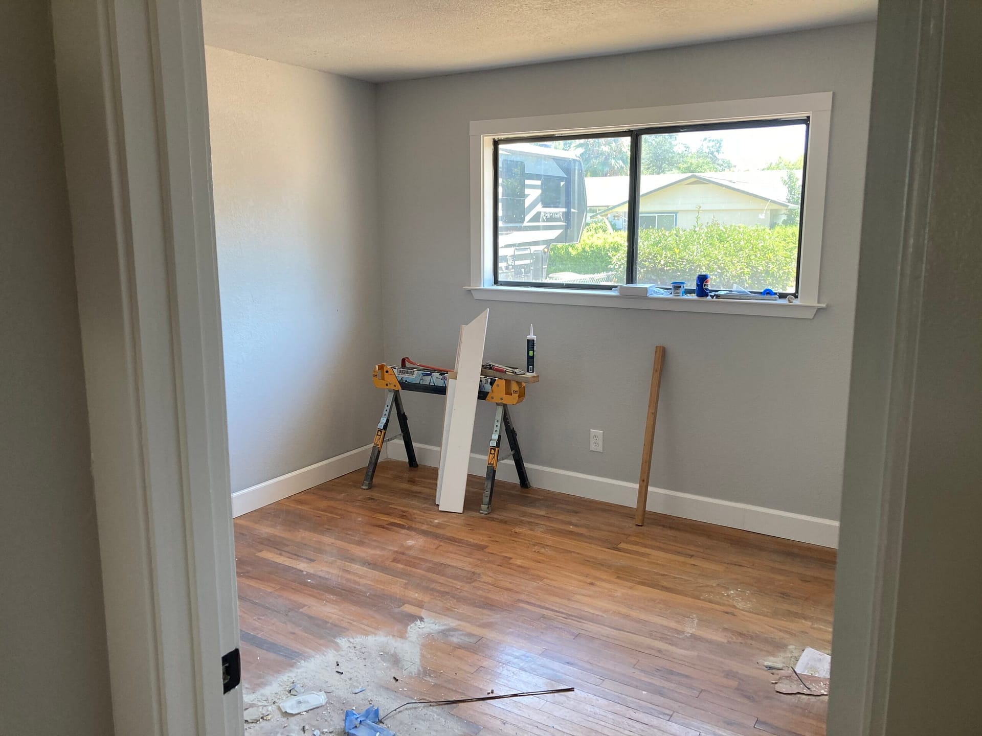 Room with a large window, gray painted walls, white baseboards, wood-look flooring and a work stand.