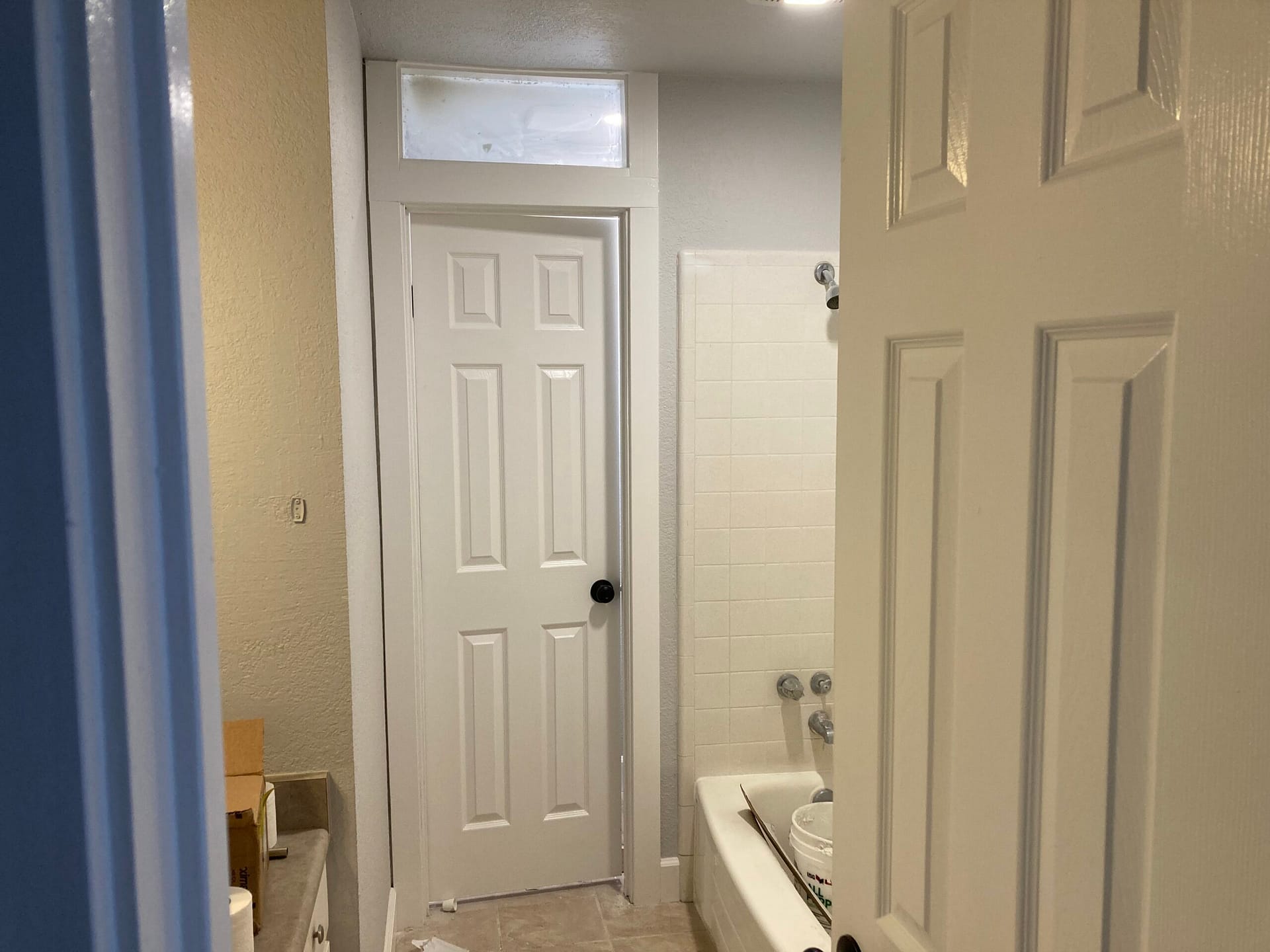 Bathroom hallway view showing white paneled doors, bathtub surround, tiled floor and plumbing fixtures during renovation.