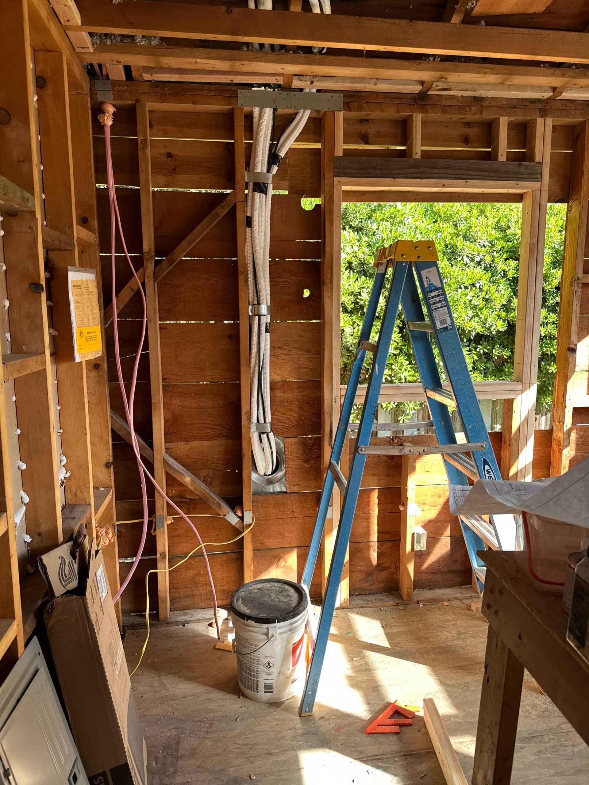 Stripped interior wall with a large framed window opening, ladder, exposed wood studs and overhead wiring.