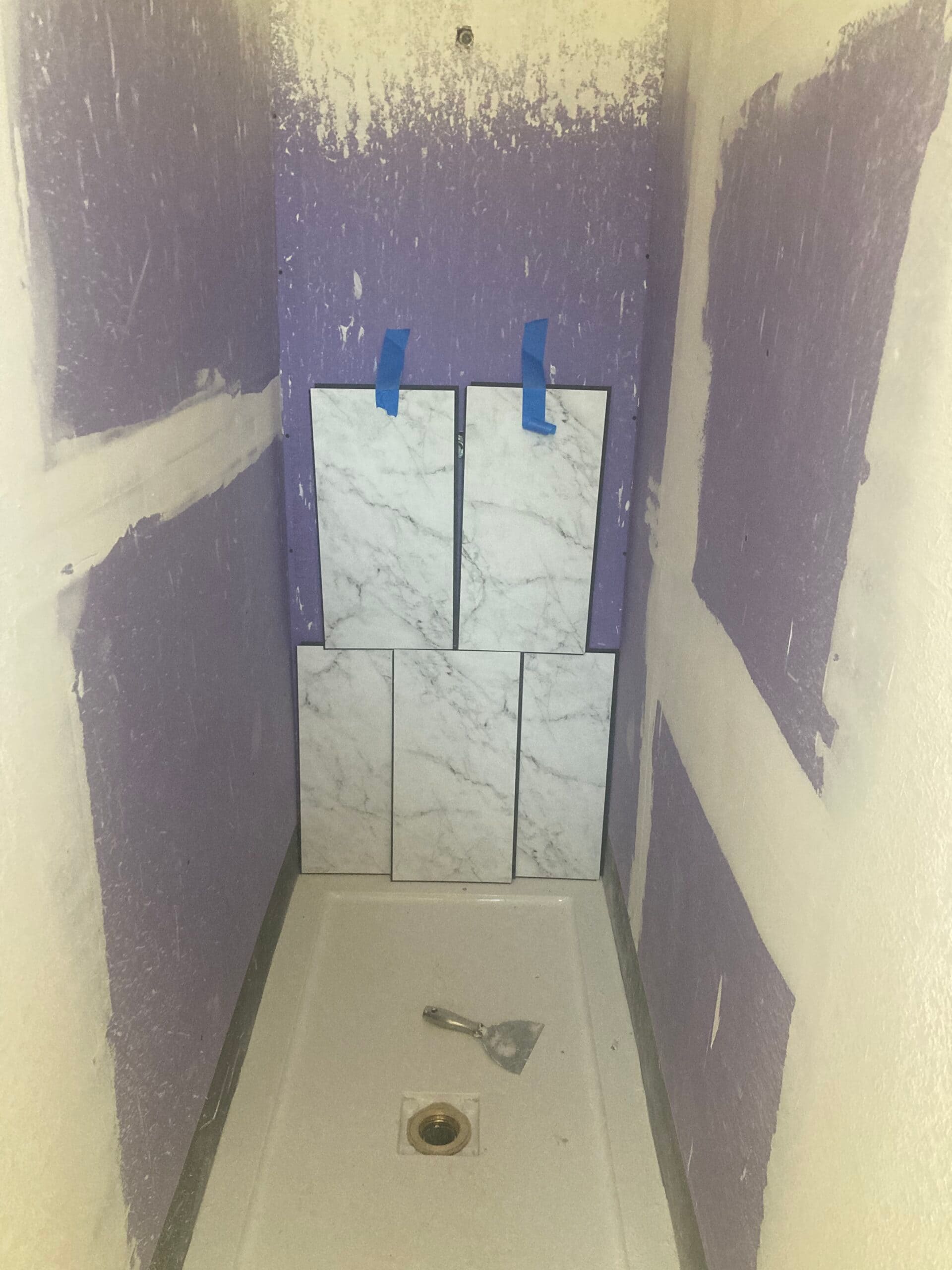 Shower enclosure with purple moisture-resistant wallboard, taped seams, a white shower base and marble-look tiles staged against the wall.