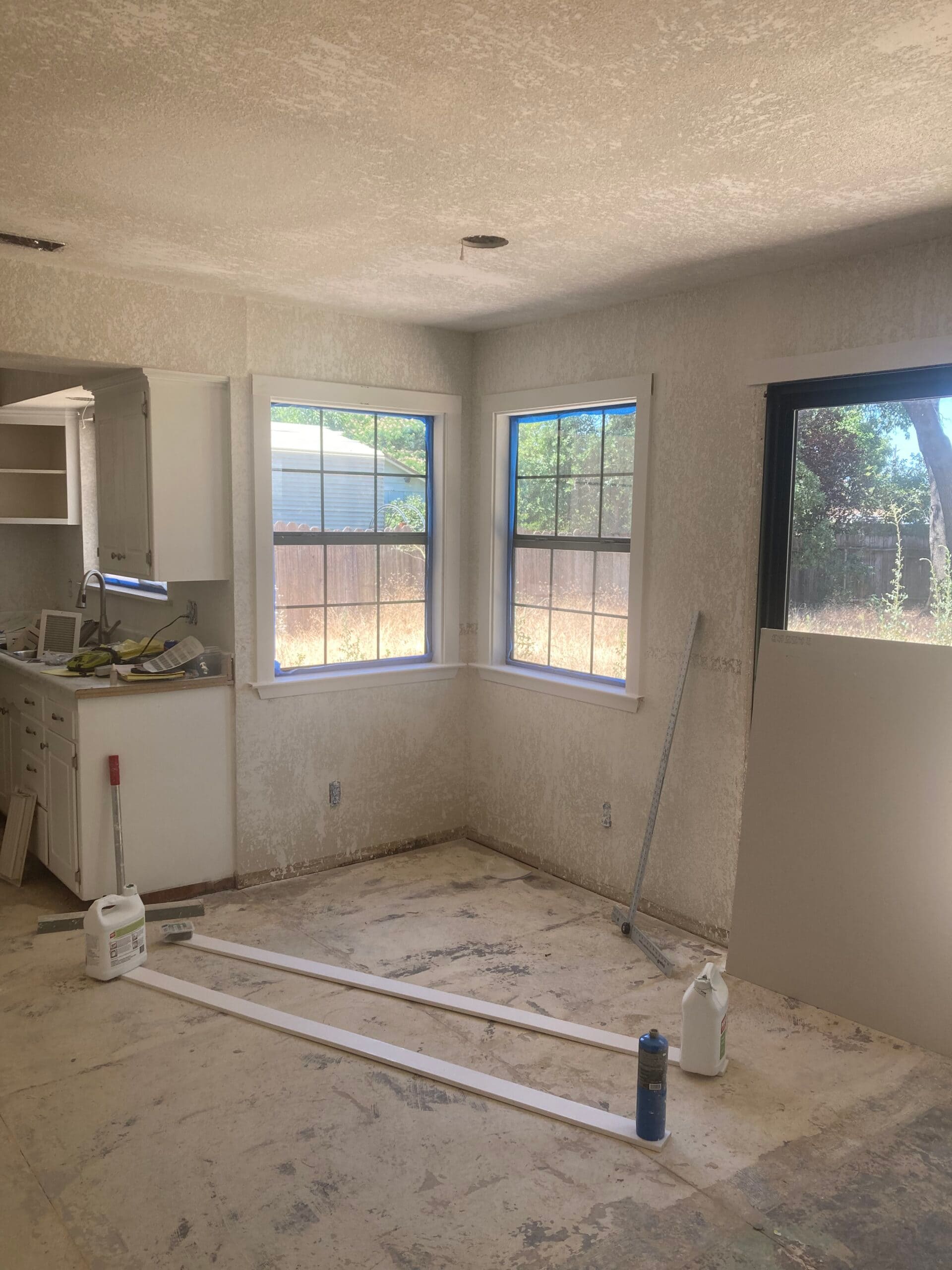 Bright living room with three windows, an opening to the kitchen and exposed floor surfaces during remodeling.