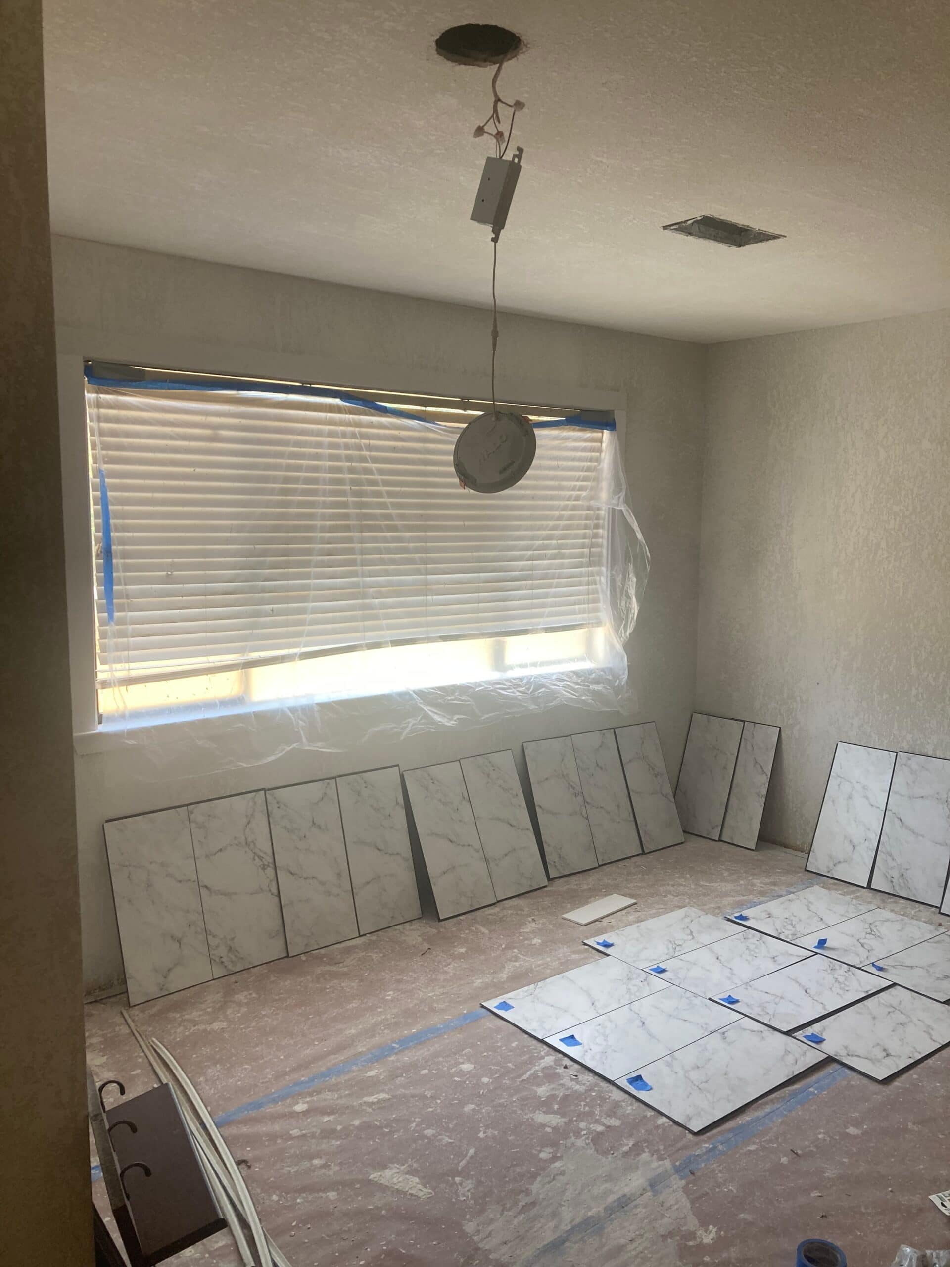 Marble-look large-format tiles laid out on an unfinished room floor, with additional tiles leaning against the wall and a hanging light.