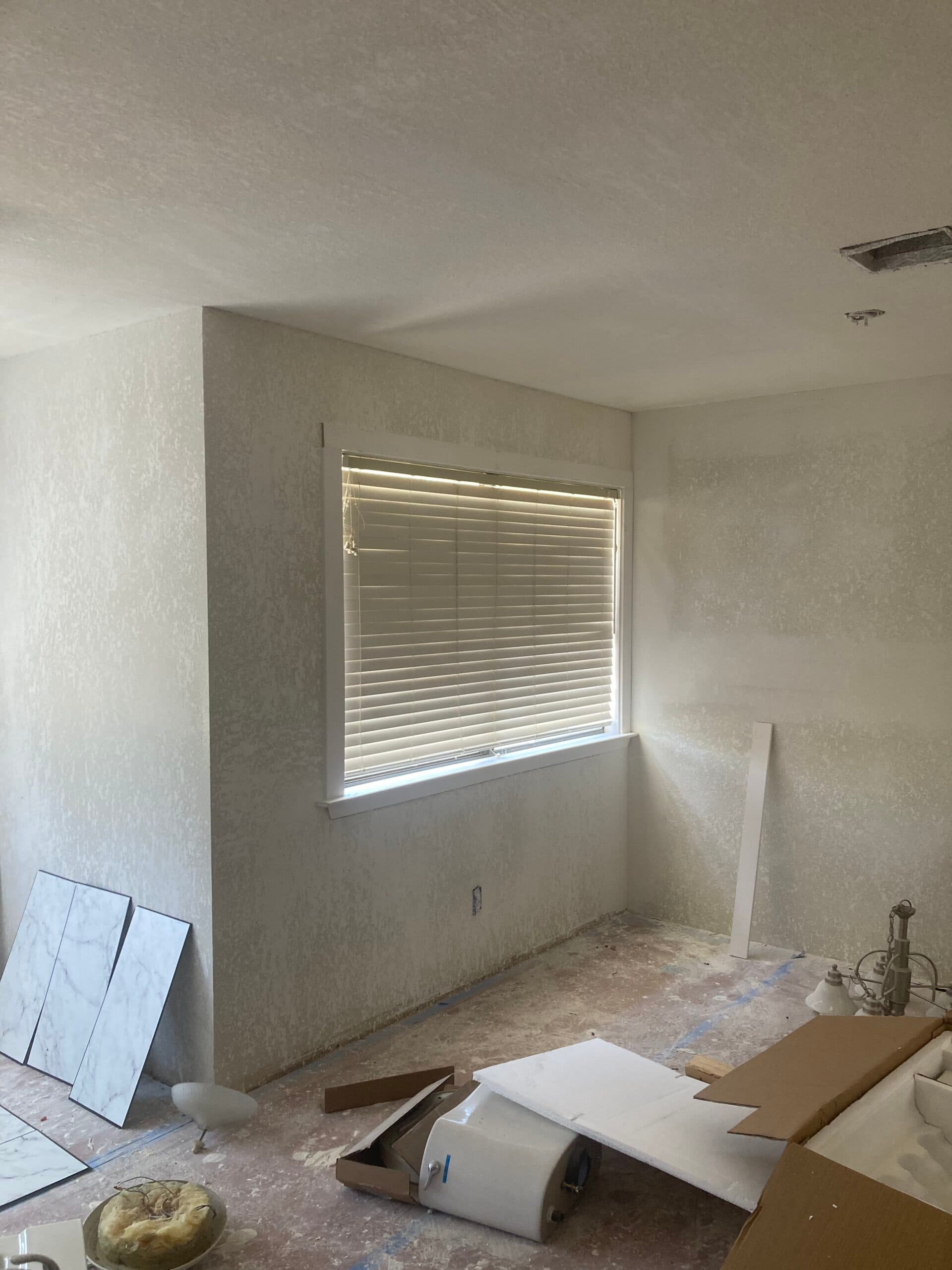 Room during remodeling with window blinds, textured walls, exposed floor and tiles or panels stacked around the edges.