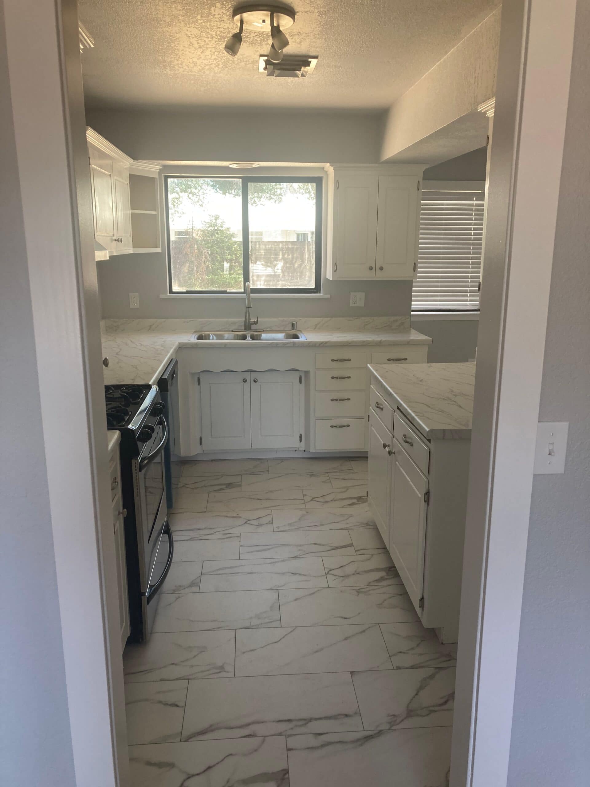 View through a kitchen doorway showing white cabinets, wide sink window, patterned floor tiles, range and counters.