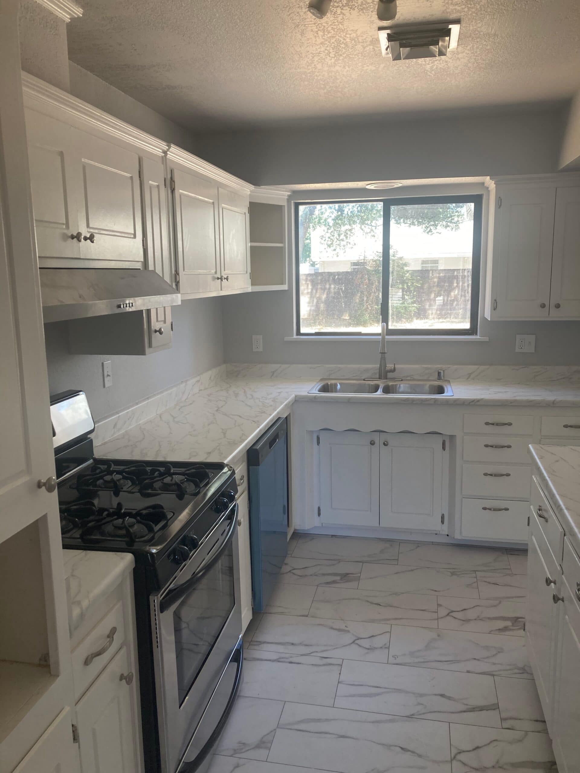 Bright white kitchen with shaker cabinets, stainless kitchen sink beneath a large window, dark stove and marble-look flooring.