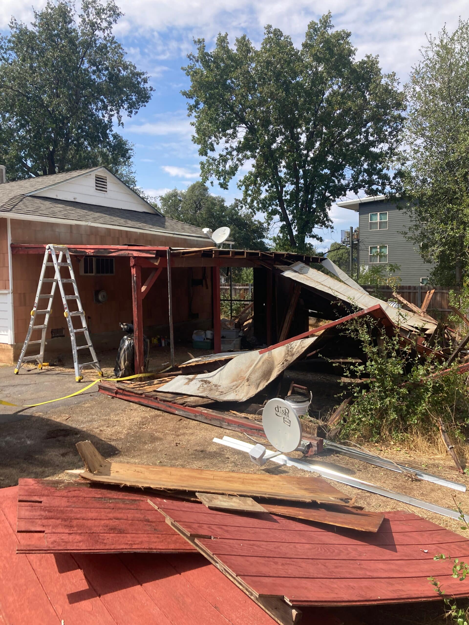 Partially collapsed exterior roof and wood structure with removed panels and debris beside a ladder during demolition.