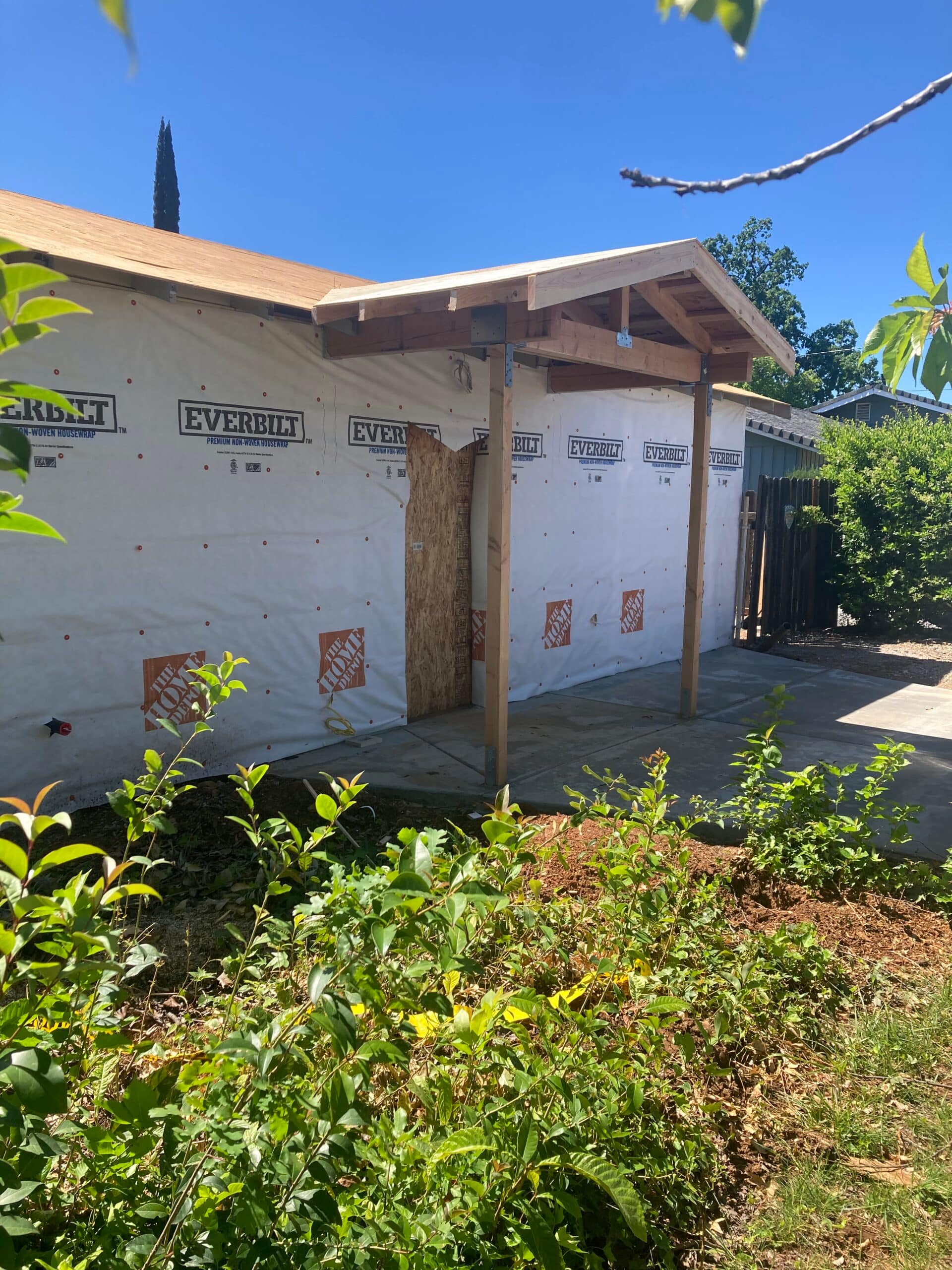 New covered entry porch construction with wood posts, gable roof framing, plywood roof sheathing, and exterior house wrap before siding installation.