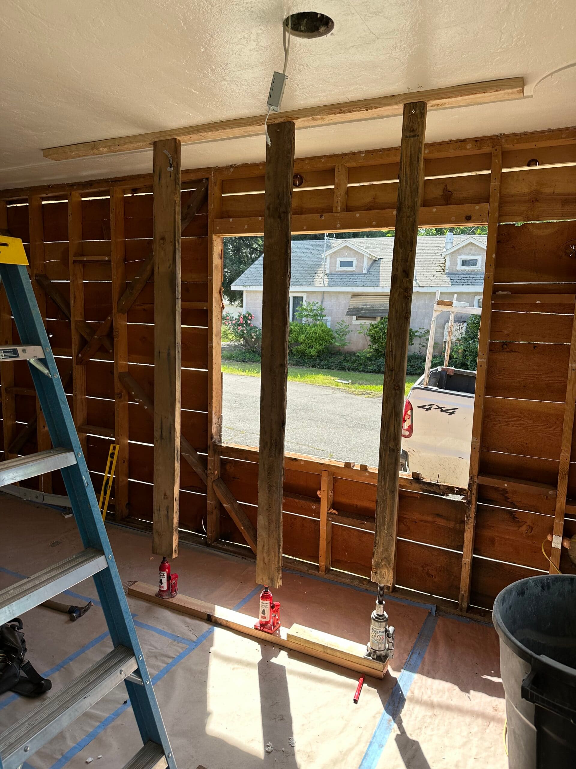 Sagging ceiling repair with three temporary wood support posts on hydraulic jacks beneath a horizontal brace, beside exposed interior wall framing.