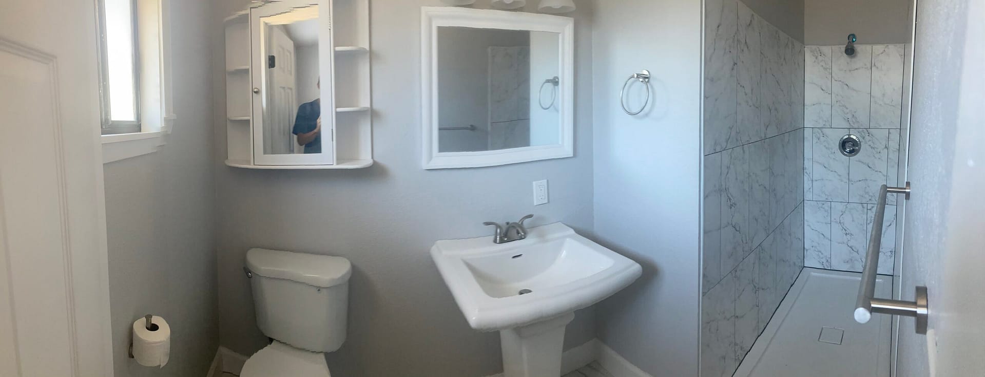 Bathroom interior with a white pedestal sink and toilet, wall-mounted cabinets and a marble-look tile shower surround.