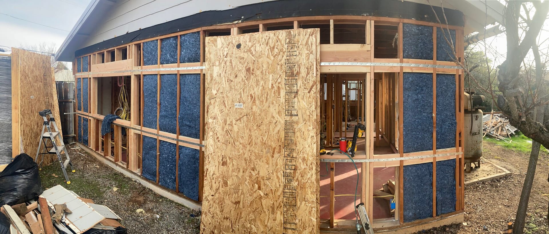 Wide exterior view of a renovation showing wall studs, insulation panels and plywood across a central door opening.