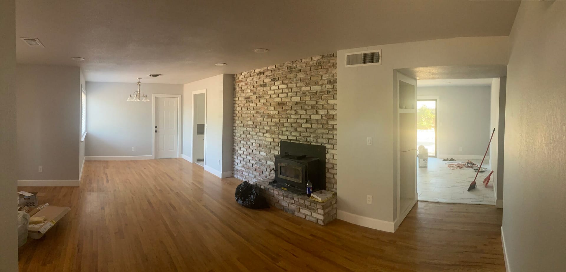 Light living room with wood-look plank floors, a pale brick fireplace and wood stove, gray walls and adjacent room openings.