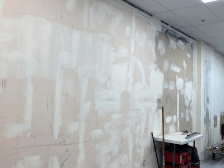 Drywall repair and finishing in progress on a commercial interior wall, with joint compound covering seams, patches and fastener holes below a suspended ceiling.