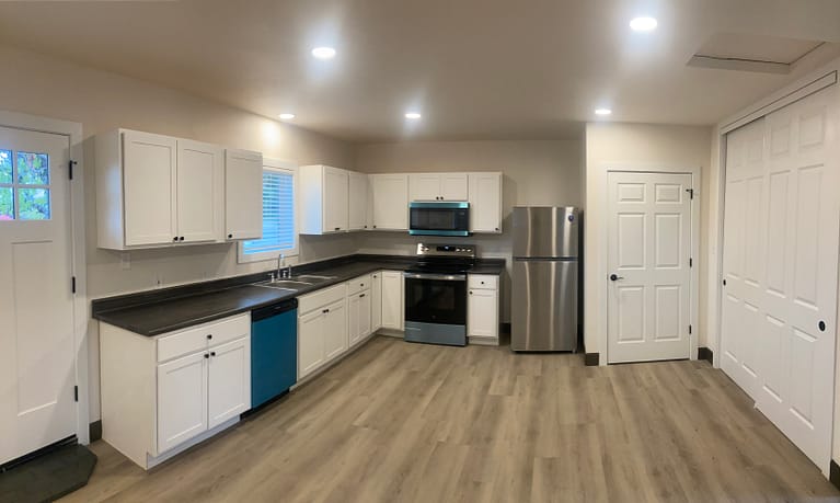 Kitchen in a Redding garage-to-ADU conversion with white shaker cabinets, dark countertops, stainless steel appliances and wood-look flooring.