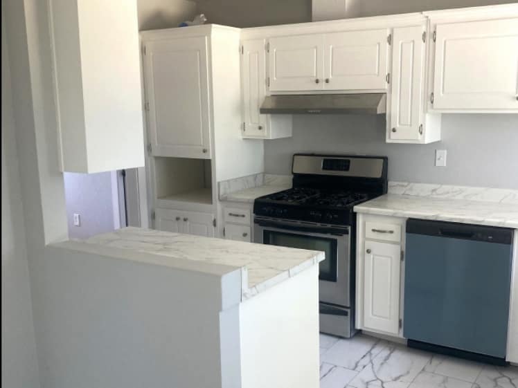 Remodeled small kitchen with white cabinets, marble-look countertops and floor tile, a peninsula counter, gas range, range hood and dishwasher.