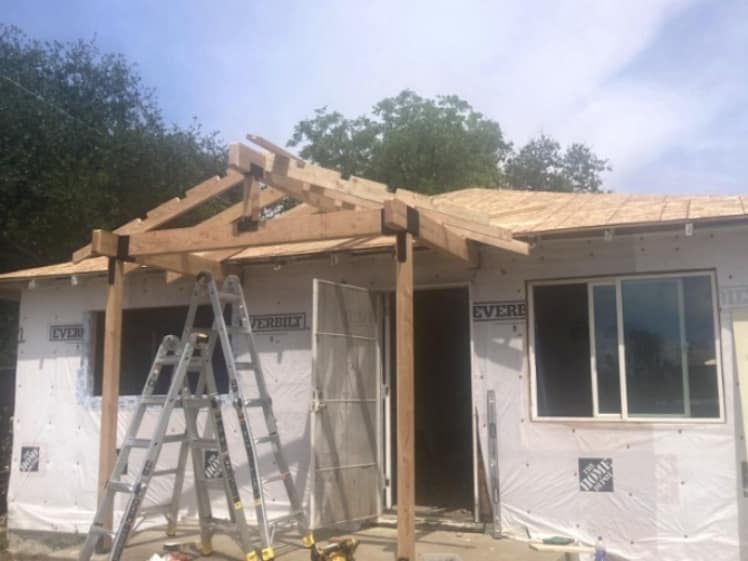 Front porch roof framing in progress with wood posts, beams and a gabled entry cover beside newly sheathed roofing and house-wrapped exterior walls.