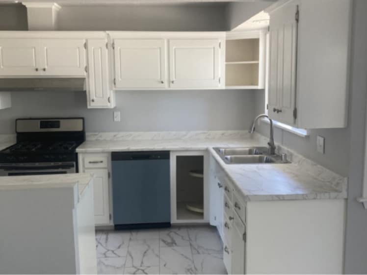 Remodeled kitchen with white cabinets, marble-look countertops and floor tile, gray walls, a double-basin sink, dishwasher and gas range.