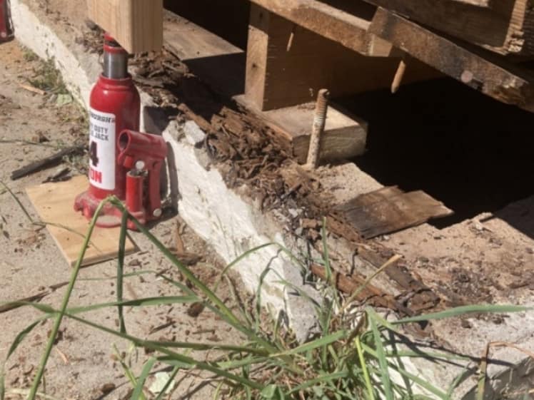Severely rotted sill plate exposed above a concrete foundation, with deteriorated wood around an anchor bolt and a hydraulic jack supporting temporary framing.
