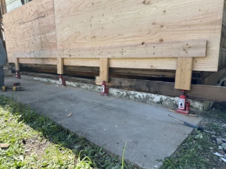 Exterior wall supported by temporary wood bracing and hydraulic jacks during dry rot repairs, with plywood sheathing above the exposed sill plate area.
